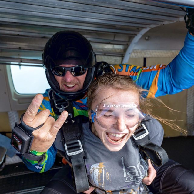 Male tandem instructor gives the horns hand symbol and female tandem instructor in a gray shirt closes her eyes before exiting the airplane.