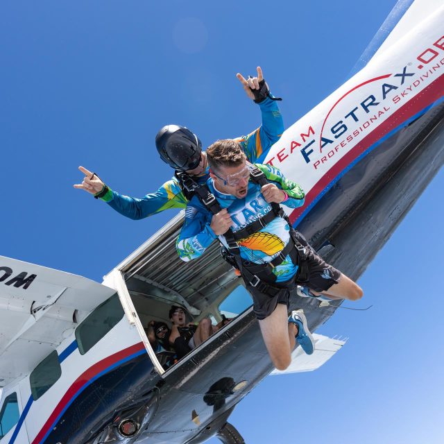 Male tandem pair exit the airplane at Start Skydiving. Instructor in black helmet gives the horns hand signal while the tandem student closes his eyes and clutches his harness.