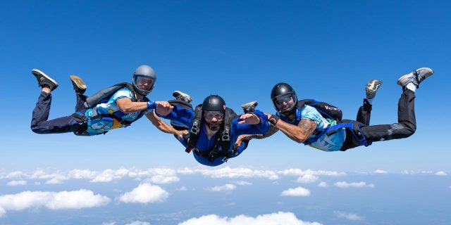 A learn to skydive AFF jumper and his two instructors, all in blue, look at the camera during freefall.