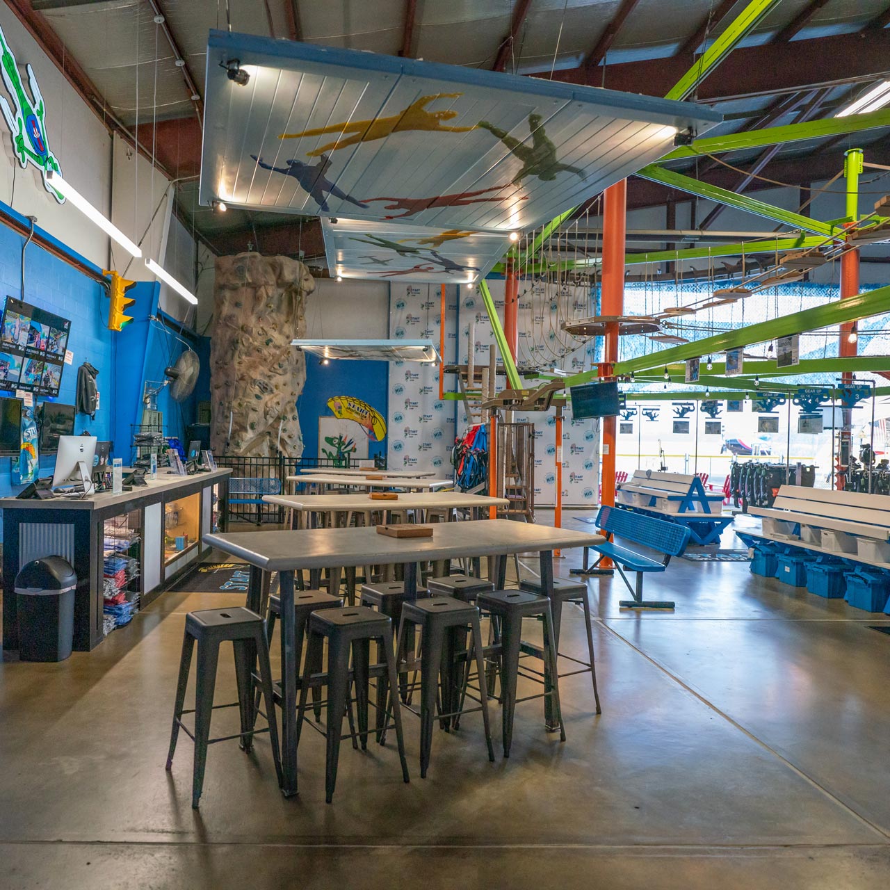 Inside Start Skydiving depicts a climbing wall, neon green ropes course, and tall tables for sitting.