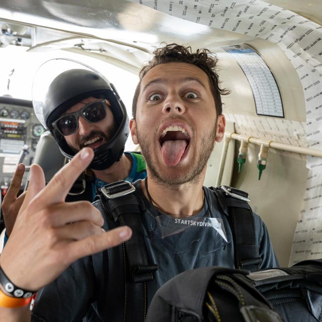Male tandem skydiver extends his tongue and gives the rock on hand signal with his skydiving instructor on the airplane.