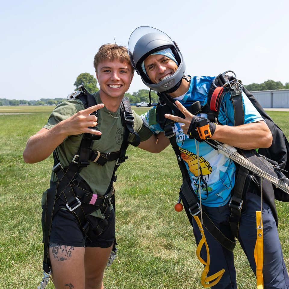 A tandem instructor and his passenger in a green shirt and black shorts pose for a photo after landing from a skydive.