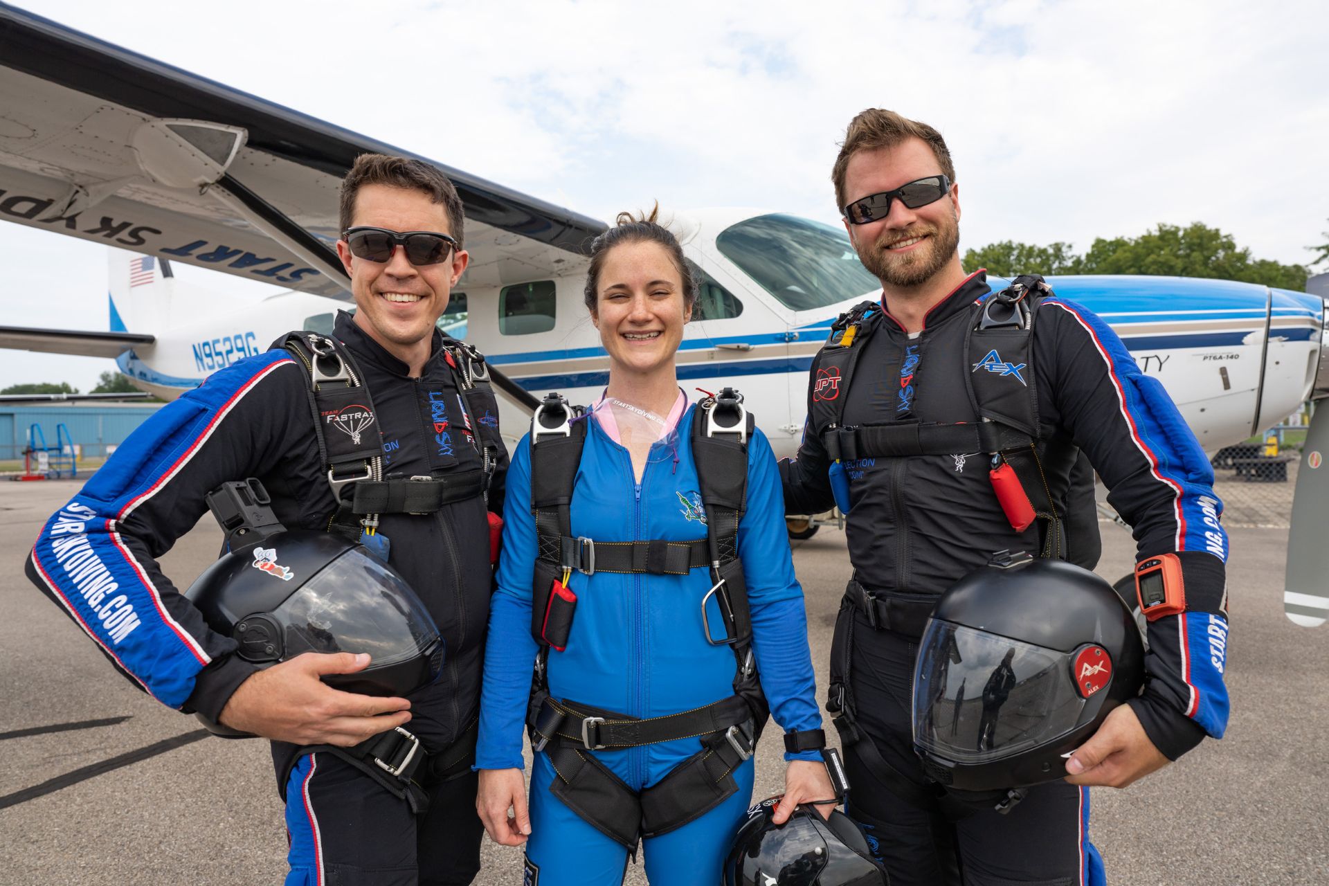 A smiling female AFF student in a blue jumpsuit stands between AFF Instructors John Hart III and Alex Hart