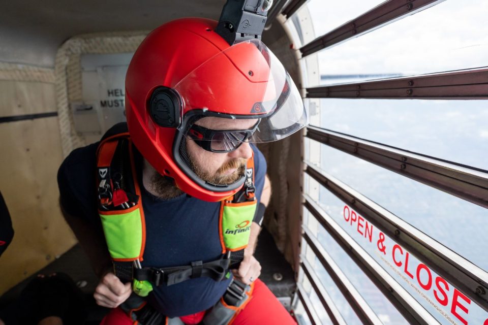 Male licensed jumper in a red helmet with a lime green and red skydiving rig gazes out the clear door of the airplane.