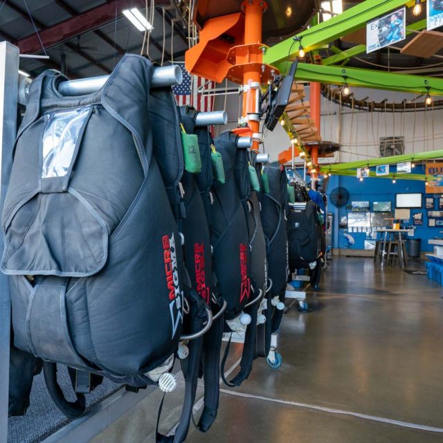 Tandem skydiving gear hangs on a rack in the foreground and the lime green and orange high ropes course lies above.