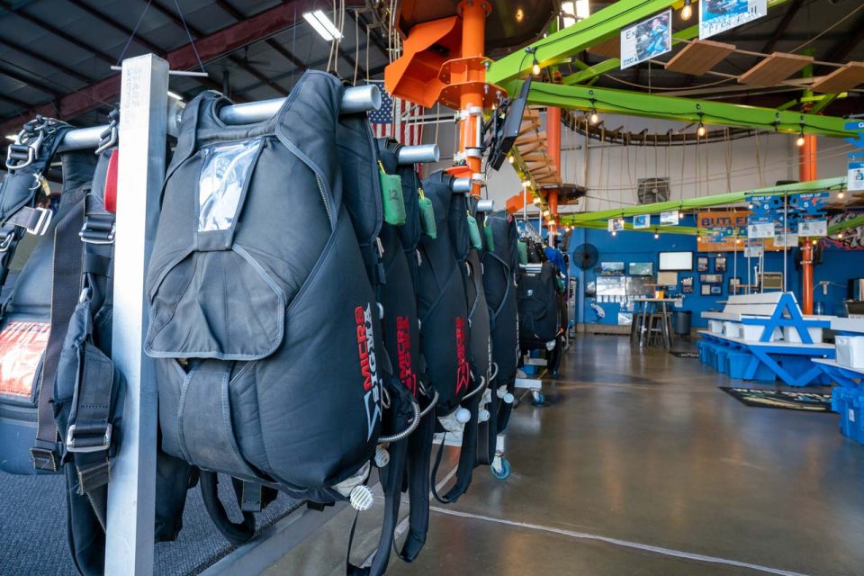 Tandem skydiving gear hangs on a rack in the foreground and the lime green and orange high ropes course lies above.