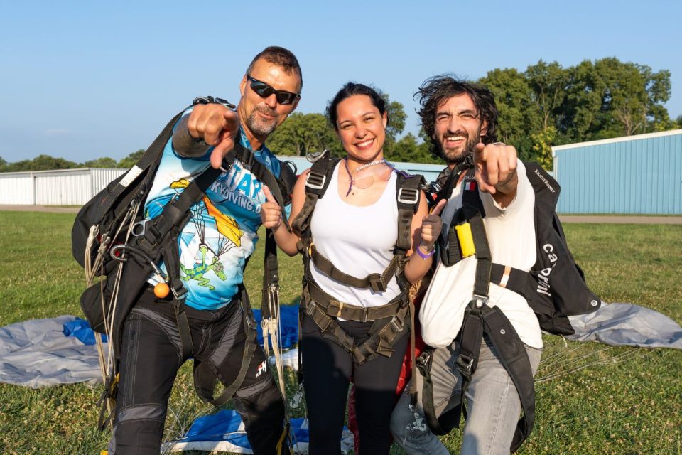 Female tandem skydiving student wearing a white tanktop and her instructor point and smile at the camera.
