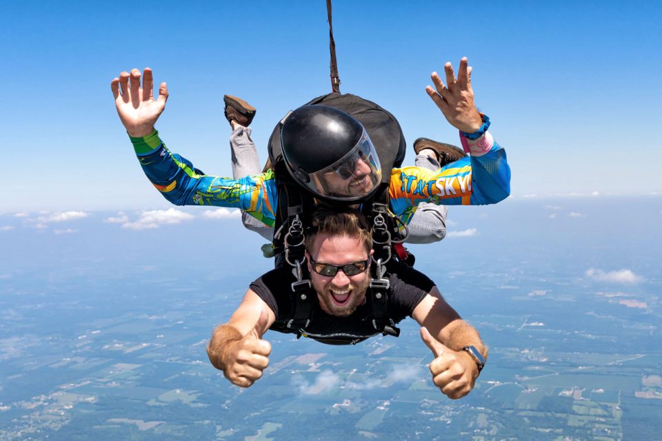 Male tandem skydiving pair giving thumbs ups and smiles during freefall.