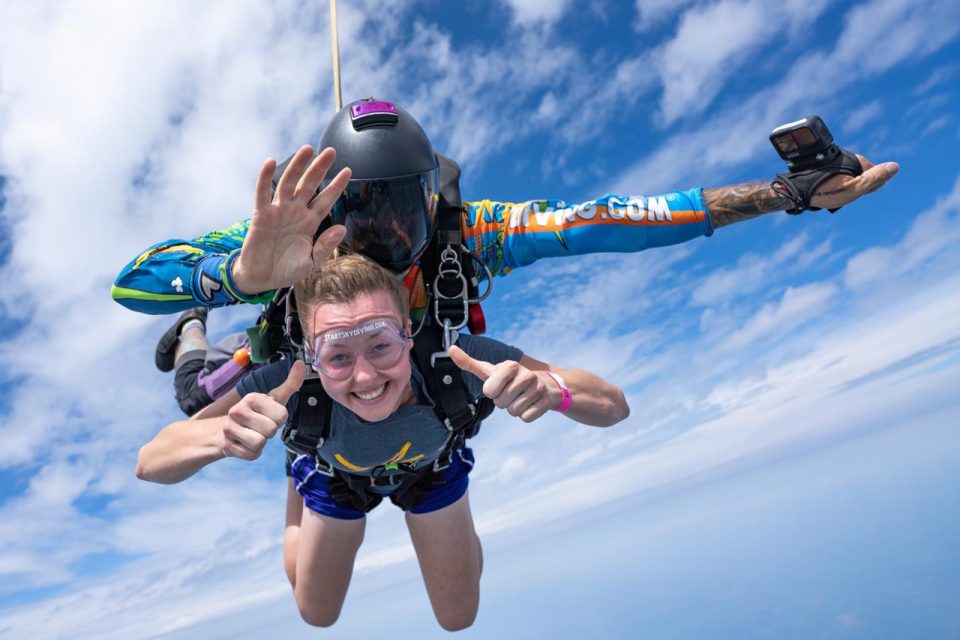 Female tandem student gives thumbs up wearing shorts and short sleeved during freefall with her instructor.