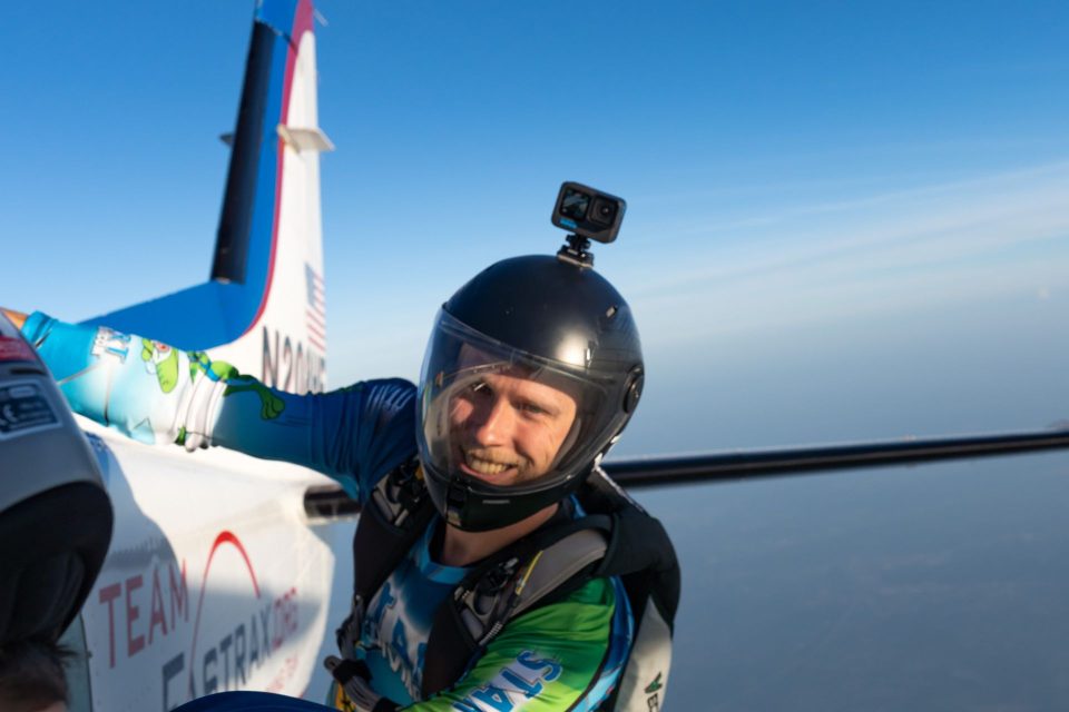 A smiling licensed skydiver wearing a gopro holds on the the aircraft from the camera step moments before exit