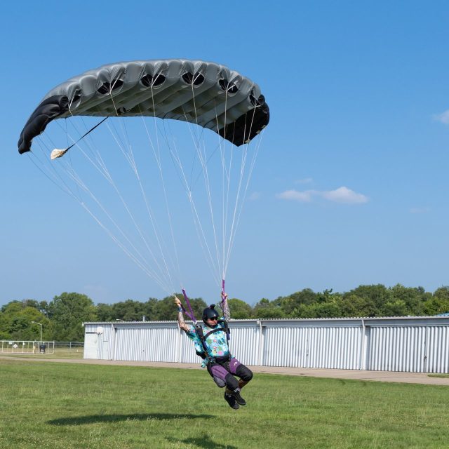A licensed skydiver flying in for landing beneath a small gray and black parachute