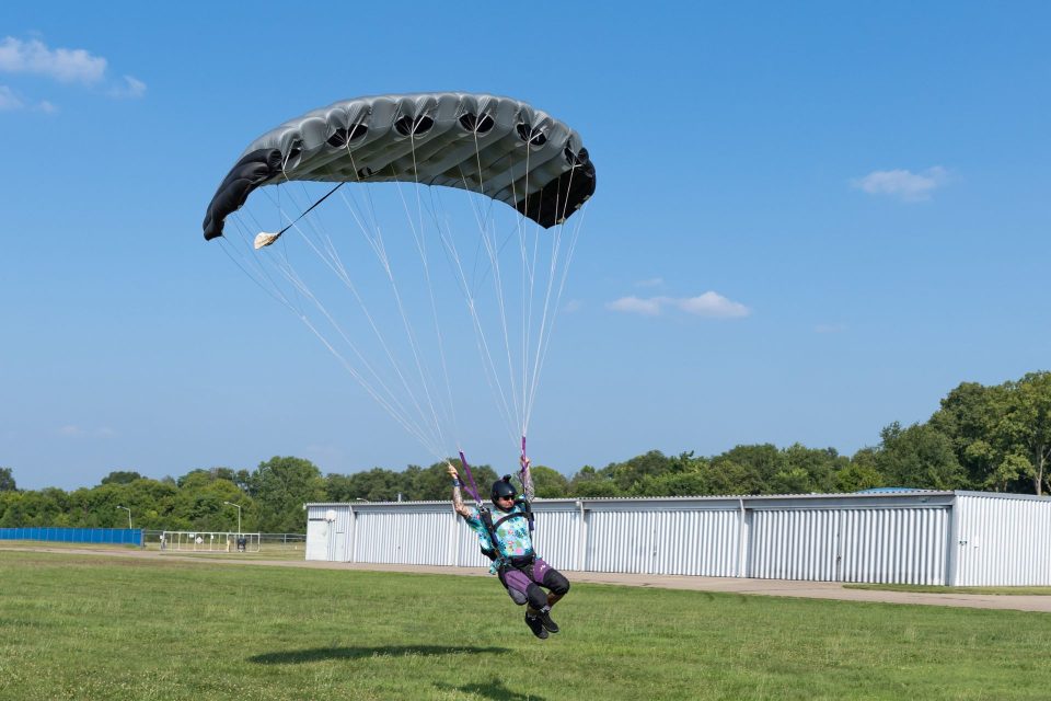 A licensed skydiver flying in for landing beneath a small gray and black parachute