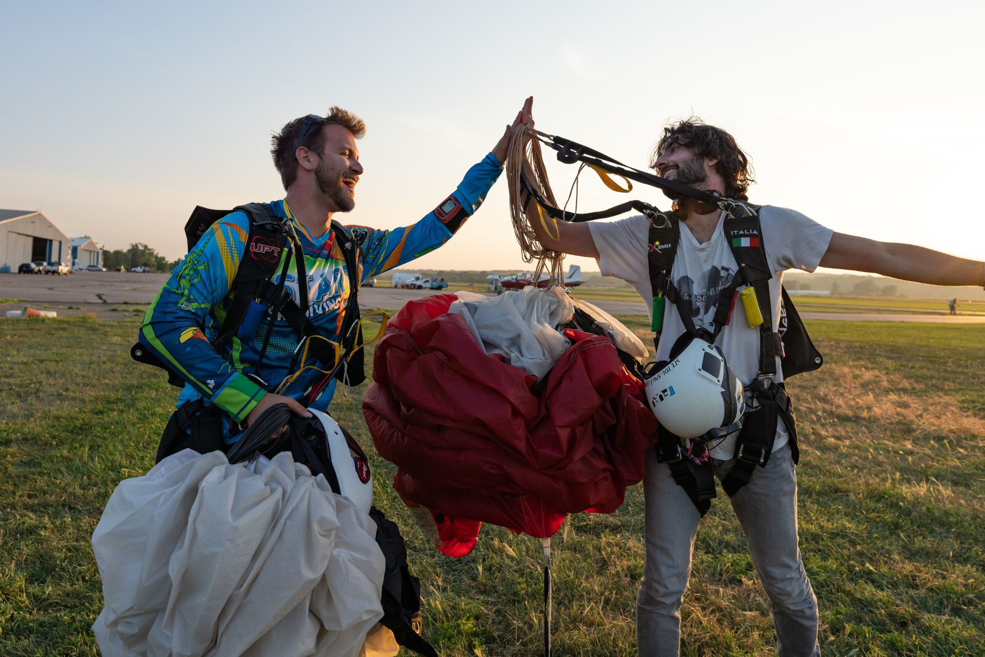 Two licensed skydivers holding their canopies high five jubilantly on the ground after landing