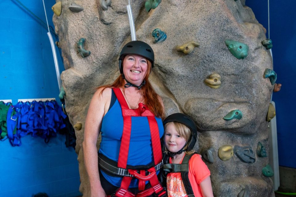 Mother and daughter stand wearing rock climbing harnesses in front of indoor rock climbing wall at Start Skydiving