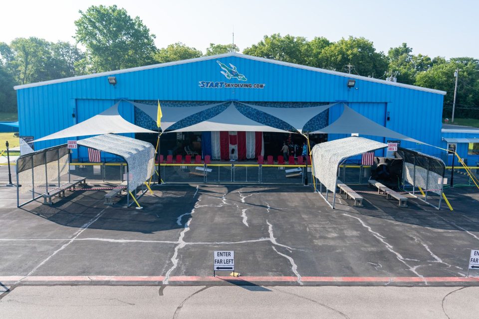 A blue metal airplane hangar with a Start Skydiving.com sign and two covered loading areas in front