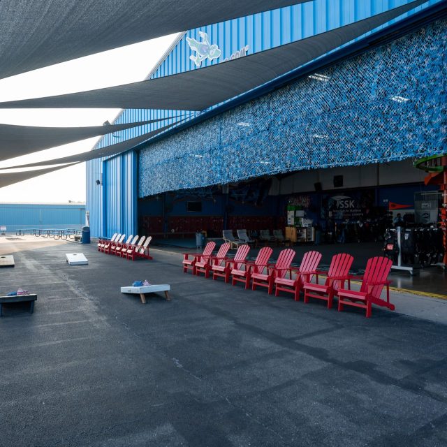 Cornhole boards and shaded outdoor seating area with red adirondack chairs at Start Skydiving