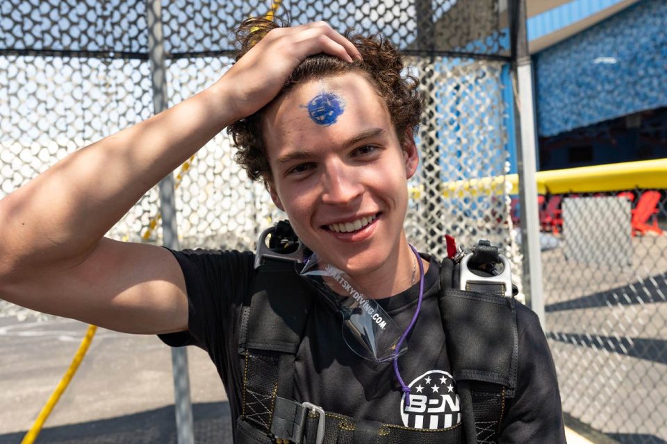 A newly licensed skydiver wearing his skydiving rig proudly pulls back his bangs to display his A license stamp on his forhead