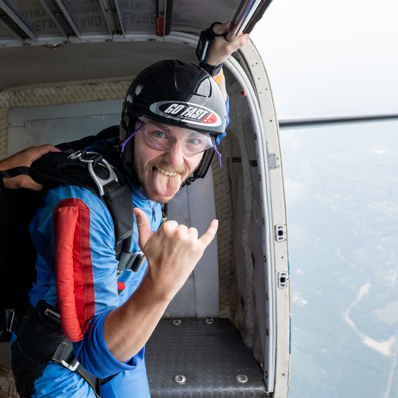 A learn to skydive AFF Student sporting a helmet with a sticker that says "Go Fast" gives the hang-loose hand signal as he prepares to exit the aircraft