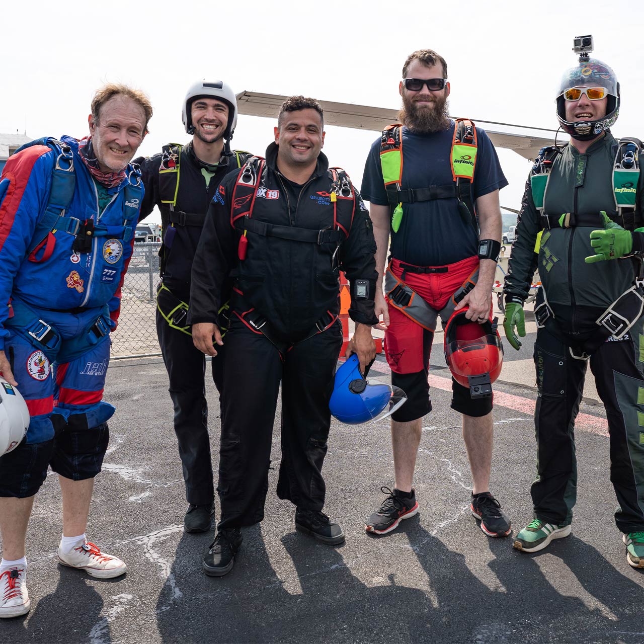 A group of licensed skydivers wearing skydiving rigs smile and pose for a photo
