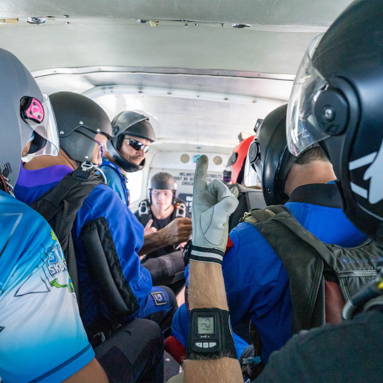 Airplane full of skydivers with a hand in the foreground giving the rock on hand symbol and wearing an altimeter that reads 12,300 feet