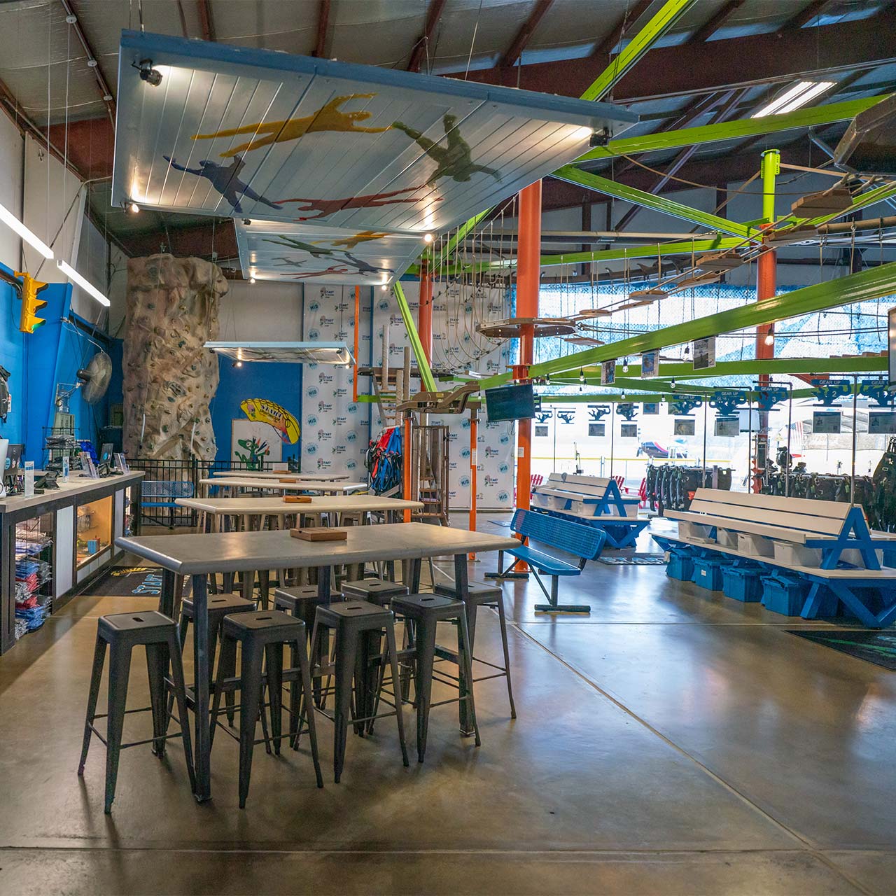 Inside Start Skydiving depicts a climbing wall, neon green ropes course, and tall tables for sitting.