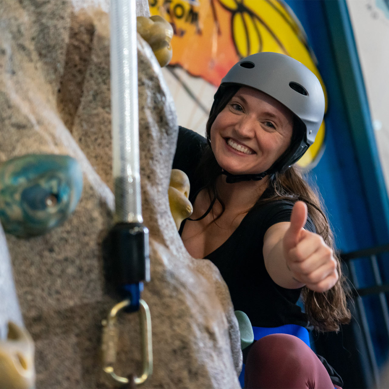 Smiling female with brunette hair gives a thumbs up while on the indoor climbing wall at Start Skydiving