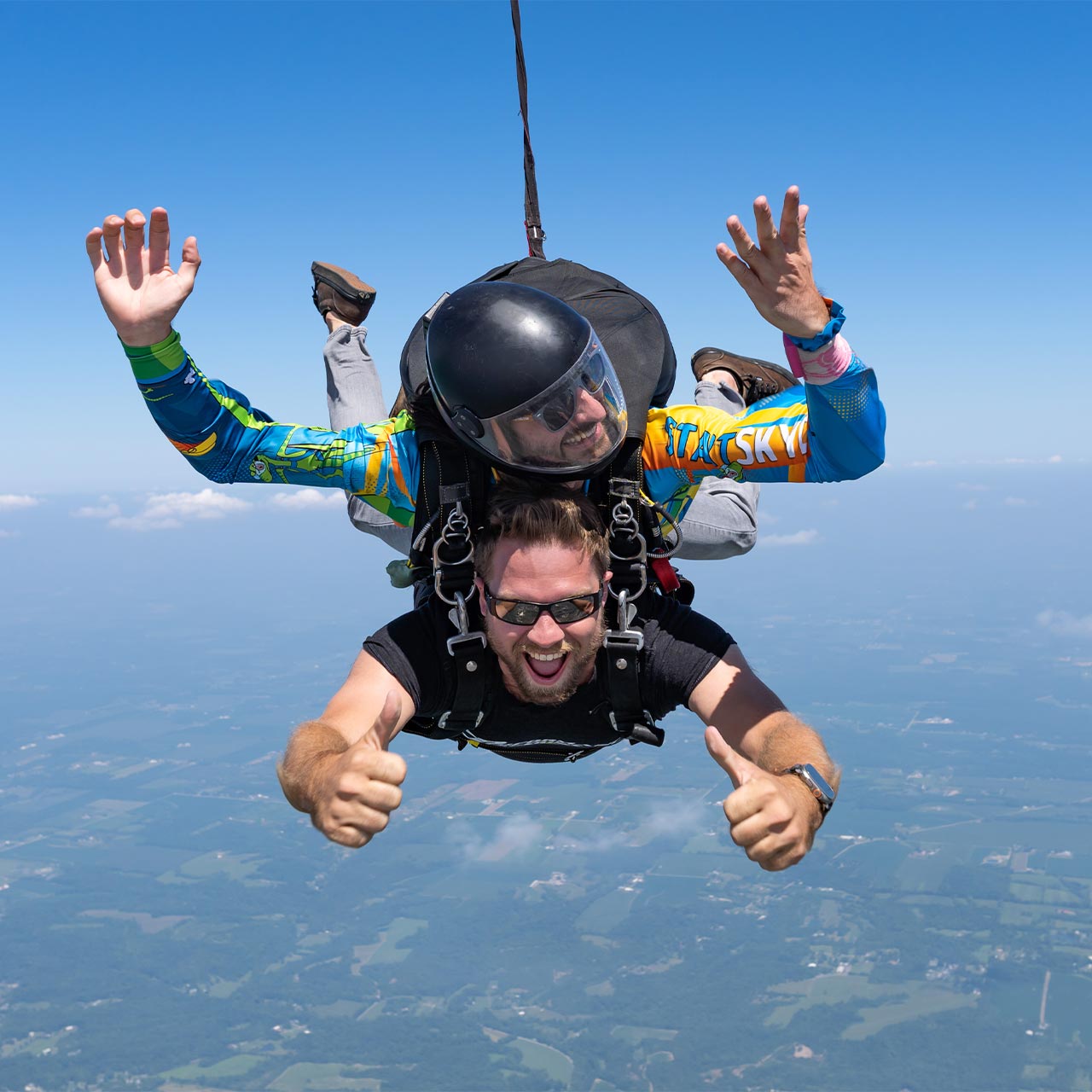 Male tandem skydiving pair giving thumbs ups and smiles during freefall.