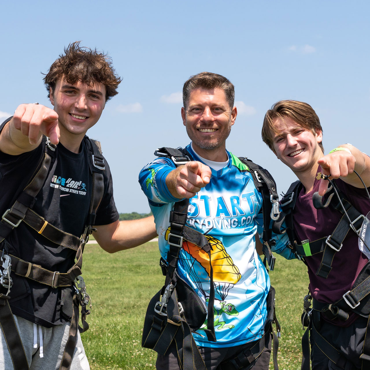 Three tandem skydivers pointing to the camera still wearing their harnesses after their jump.