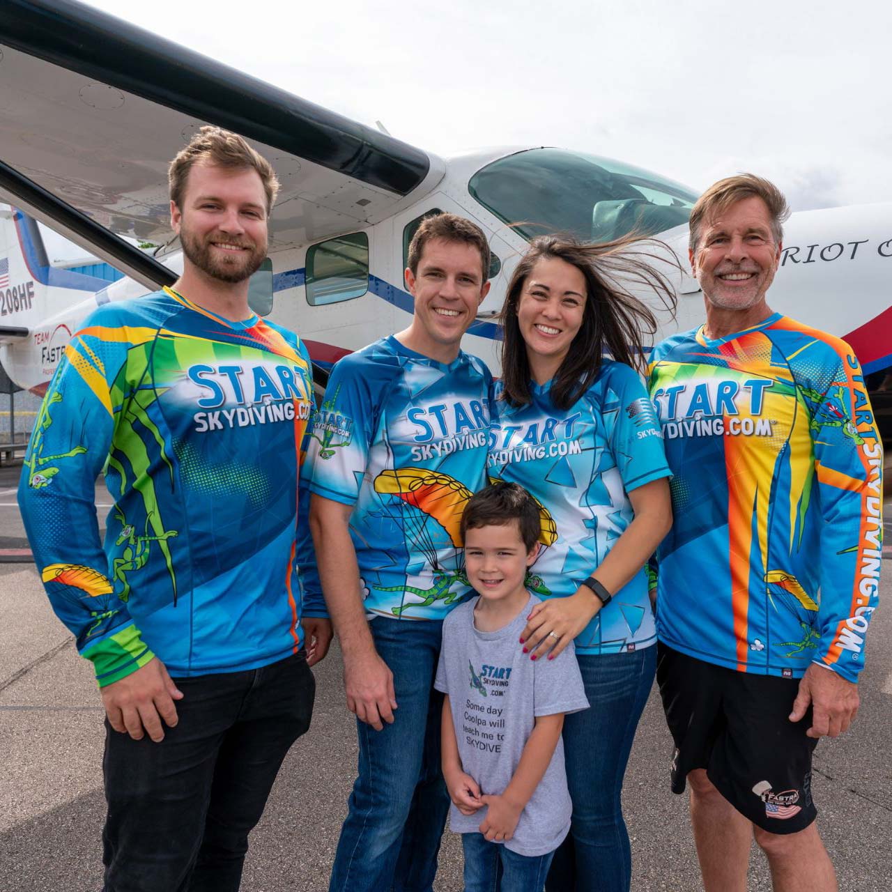 Alex Hart, John Hart II, married couple John Hart III and Lisa Hart, and their young son smile wearing Start Skydiving team jerseys in front of the Patriot One Aircraft.