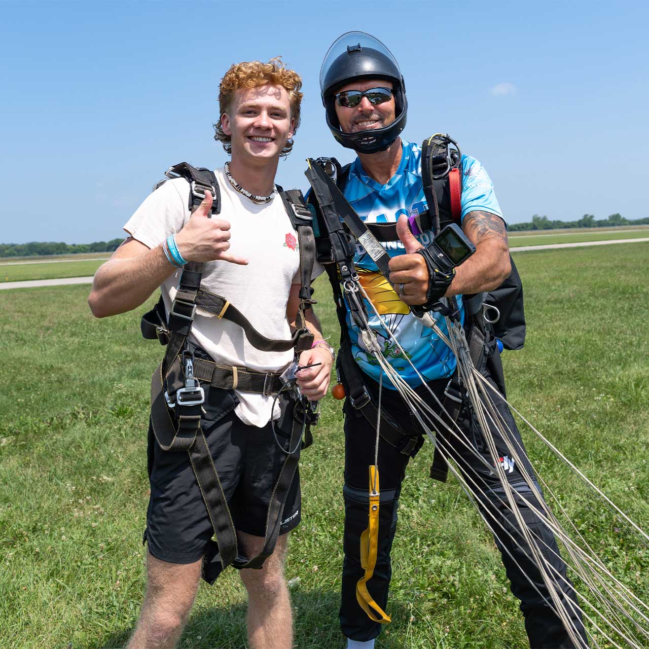 male tandem student with red hair wearing a white t-shirt posing after landing giving the hang loose sign as the tandem instructor gives a thumbs up