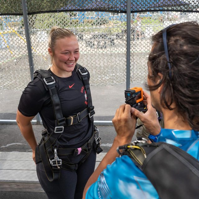 A blonde female tandem student wearing a skydiving harness being interviewed on film by her tandem instructor